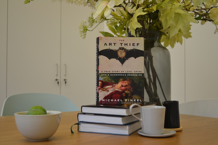 Op een tafel ligt een stapel boeken. Er staat een kommetje fruit, een kopje en bos bloemen naast.