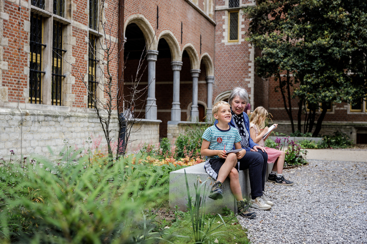 Twee kinderen en een volwassene zitten op een bankje in de tuin. Op de achtergrond is het stadspaleis met pilaren te zien.