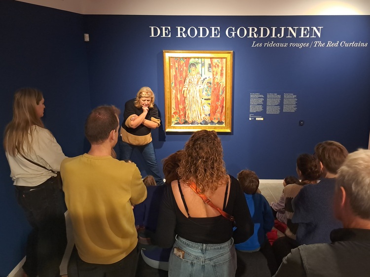 Een gids praat tegen een groep naast een schilderij dat aan een blauwe muur hangt.