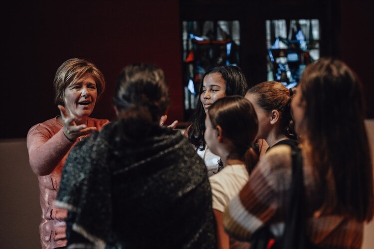 Een groep tieners staat in een museumzaal en praten met een lachende vrouw.