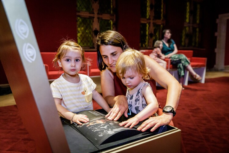 Een vrouw met twee kleine kindjes opent een gouden kubus in een museumzaal.