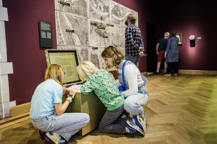 Deux enfants et un adulte regardent à l'intérieur d'un cube doré situé dans un hall de musée.