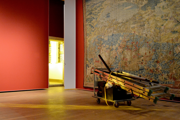 Het licht van binnen in een museumzaal met rode muren en houten vloer. Er hangt een groot wandtapijt aan de muur. Centraal staat een kar met werkmateriaal, waaronder een ladder.